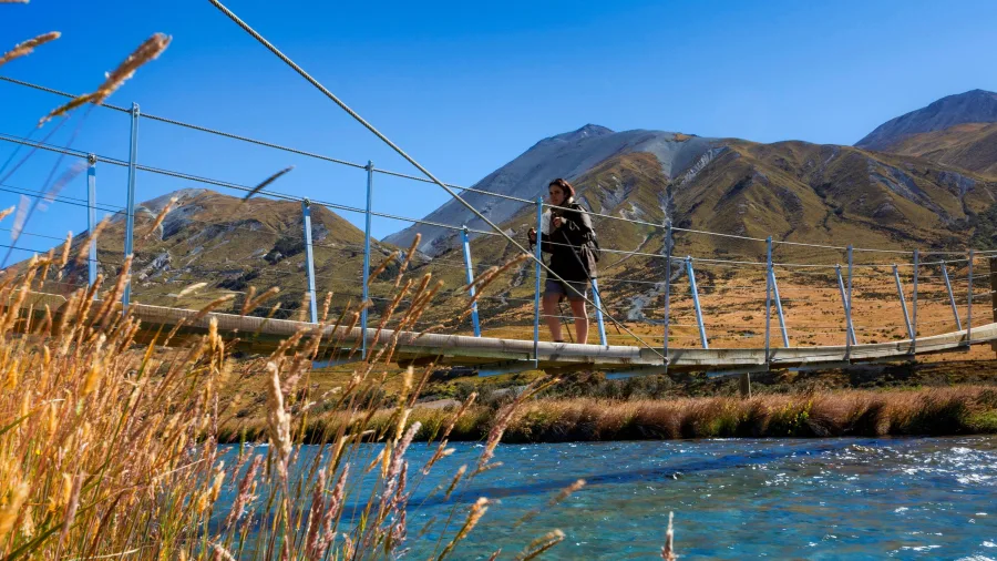 Hiker crossing the Mt Sunday footbridge over a clear river with Lake Clearwater and mountain ranges in the Ashburton Lakes district, Mid Canterbury, New Zealand