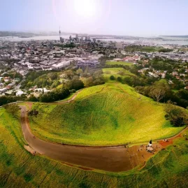Aerial view of Mount Eden volcanic crater with visitors on walking paths during golden hour, Auckland city visible in background.