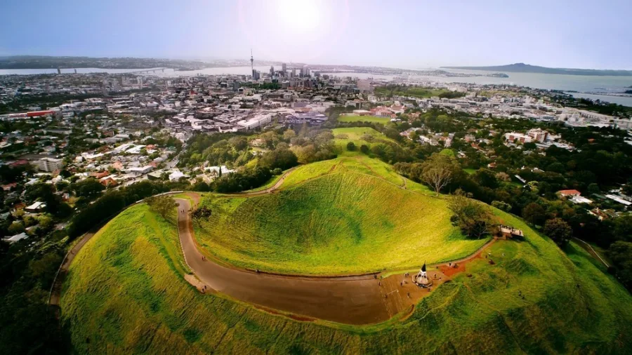Aerial view of Mount Eden volcanic crater with visitors on walking paths during golden hour, Auckland city visible in background.