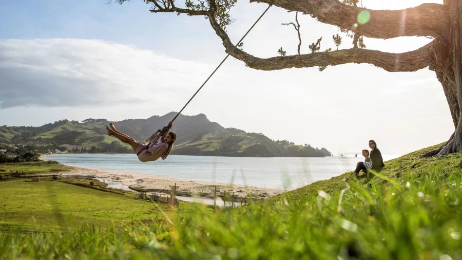 Child swinging from a tree overlooking Opito Bay with views to the Mercury Islands