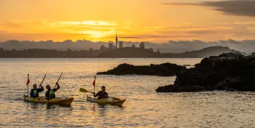 paddlers kayaking at sunset on Waitemata Harbour with Rangitoto Island and Auckland skyline in the distance