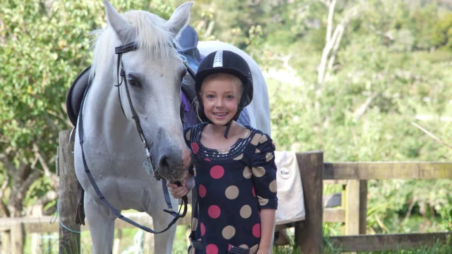 Child in a polka dot dress wearing a riding helmet stands beside a white horse in the scenic countryside of Matakana near Auckland, New Zealand