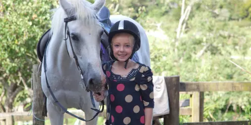 Child in a polka dot dress wearing a riding helmet stands beside a white horse in the scenic countryside of Matakana near Auckland, New Zealand