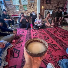 A group sits in a circle on a patterned mat for a kava ceremony, with a hand holding a bowl of kava in the foreground at Four Shells Kava Lounge in Auckland.