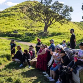 Tour group seated on logs, listening to a guide at Māngere Mountain in Auckland, surrounded by lush green hills and native trees.