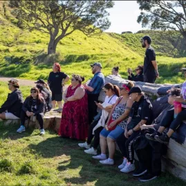 A small group of people sits and stands on large logs in a grassy, sunlit area during a guided tour of Māngere Mountain in Auckland.