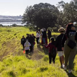 Tour group walks along a grassy path on Māngere Mountain in Auckland, enjoying scenic views of harbours and lush landscape during a cultural experience.