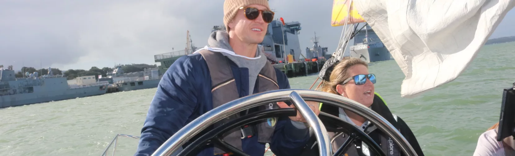 Person steering a yacht on Auckland Harbour with a crew member beside them, under cloudy skies.
