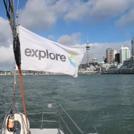 An "Explore" branded flag waving on a sailing boat in Auckland Harbour with the city skyline in the background