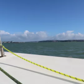A scenic view of Waitematā Harbour from the deck of a sailing yacht, with blue skies and distant land in the background.