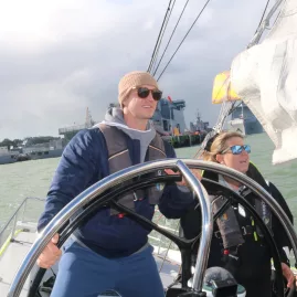 Person steering a yacht on Auckland Harbour with a crew member beside them, under cloudy skies.