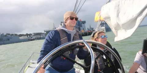 Person steering a yacht on Auckland Harbour with a crew member beside them, under cloudy skies.