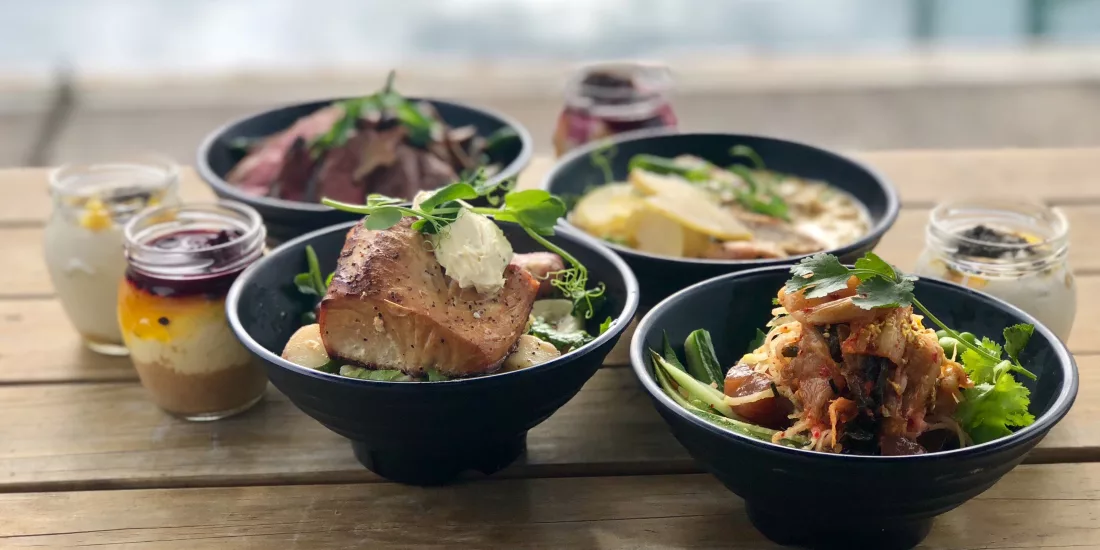 A selection of gourmet meals served in black bowls on a wooden deck, with the ocean in the background.