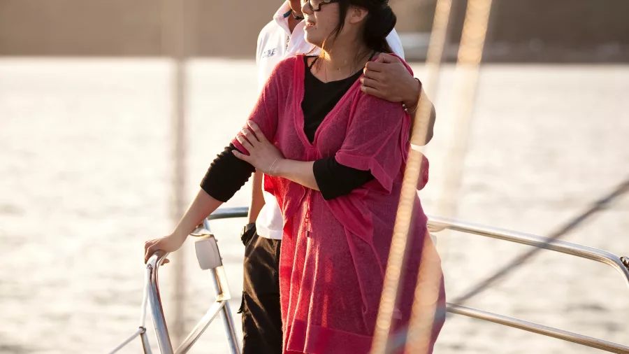 A couple embraces at the bow of a yacht, gazing at the horizon as the sun sets over calm waters.