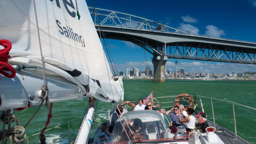 A sailing yacht with passengers enjoying the view while cruising under the Auckland Harbour Bridge with the city skyline in the background.