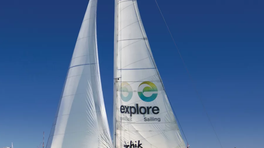 A sailing yacht with Explore Sailing branding gliding across Waitematā Harbour with Auckland’s cityscape in the background