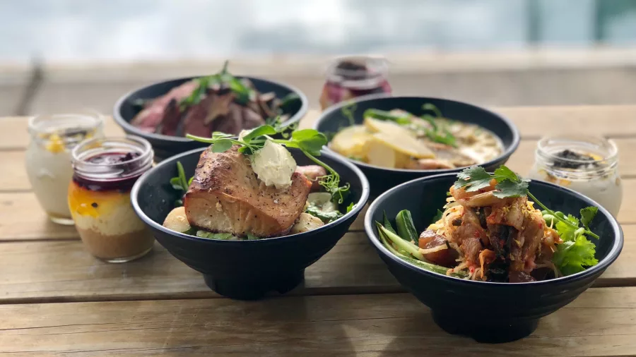 A selection of gourmet meals served in black bowls on a wooden deck, with the ocean in the background.