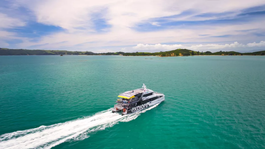 A ferry cruising through the turquoise waters of the Bay of Islands, leaving a white trail behind under a partly cloudy sky.