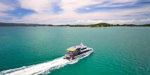 A ferry cruising through the turquoise waters of the Bay of Islands, leaving a white trail behind under a partly cloudy sky.
