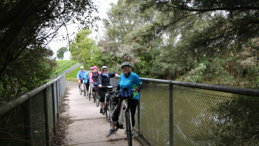Cyclists pushing bikes across a narrow bridge over a quiet waterway in Auckland