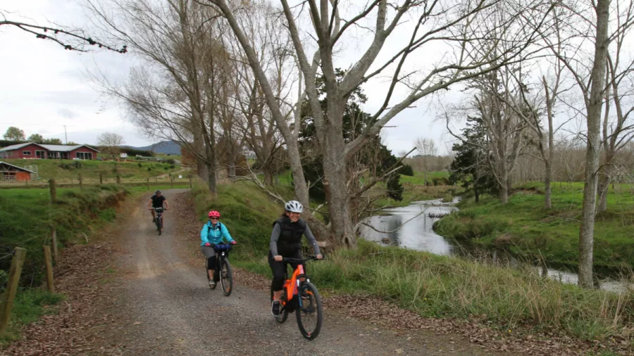 Cyclists riding e-bikes beside a stream on a gravel path through rural farmland