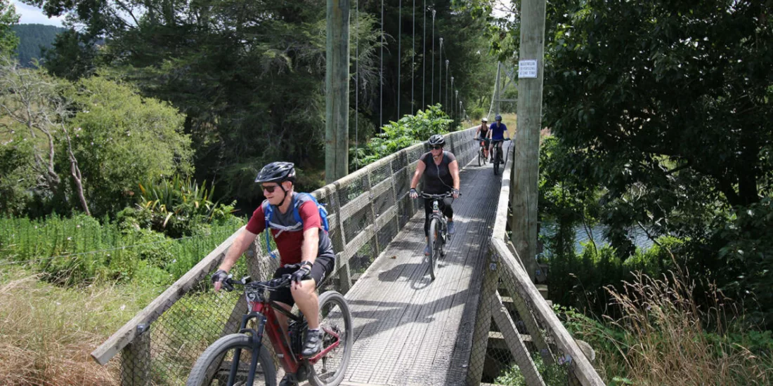 Group of cyclists riding across a wooden suspension bridge in Karangahake Gorge