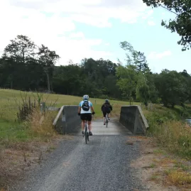 Cyclists crossing a low wooden bridge on a gravel trail in rural Karangahake Gorge