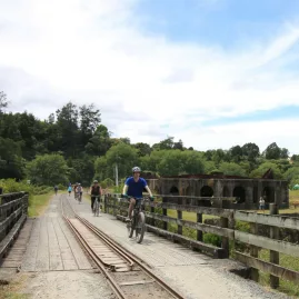 Cyclists riding over a bridge with old mining buildings visible in Karangahake Gorge