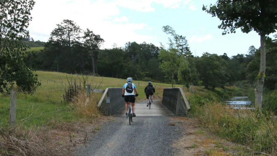 Cyclists crossing a low wooden bridge on a gravel trail in rural Karangahake Gorge