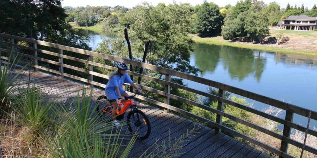 Young cyclist riding an orange bike along a wooden riverside boardwalk in Waikato