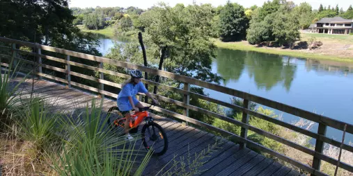 Young cyclist riding an orange bike along a wooden riverside boardwalk in Waikato