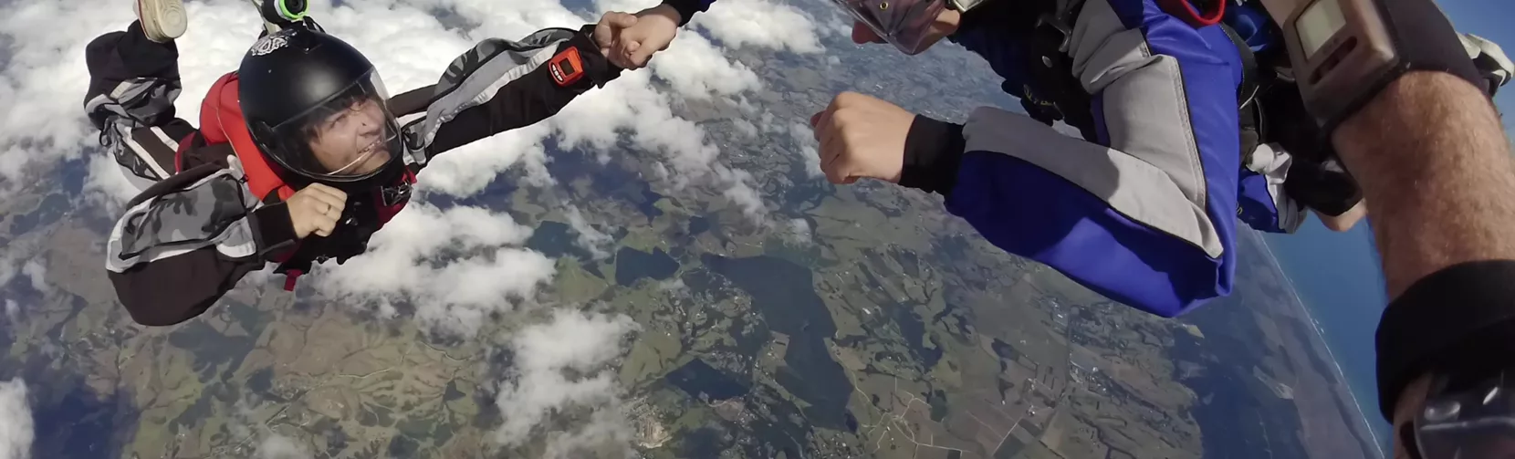 Tandem skydivers in mid-air sharing a hand grab during freefall above the Auckland region, with coastal and rural views below.