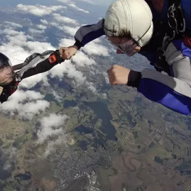 Tandem skydivers in mid-air sharing a hand grab during freefall above the Auckland region, with coastal and rural views below.