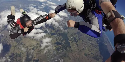 Tandem skydivers in mid-air sharing a hand grab during freefall above the Auckland region, with coastal and rural views below.
