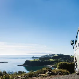 Tour van parked at the Palm Beach Lookout on Waiheke Island, overlooking a breathtaking coastal view with lush greenery and clear blue skies.