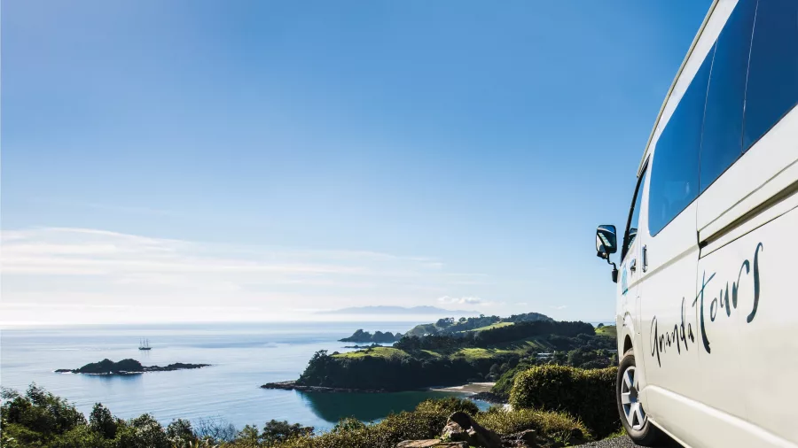Tour van parked at the Palm Beach Lookout on Waiheke Island, overlooking a breathtaking coastal view with lush greenery and clear blue skies.