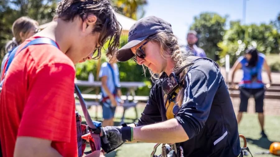 EcoZip Waiheke guide assists guest with harness and safety gear outdoors before zipline adventure, with green trees and other visitors in the background