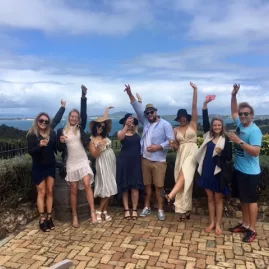 Group of people standing outdoors at a Waiheke vineyard, raising glasses and celebrating during a Premium Wine Tour with scenic coastal views in the background