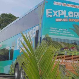 Waiheke Explorer Hop-On Hop-Off double-decker bus parked near greenery, promoting island tours and frequent stops for beaches, vineyards, restaurants, and walking tracks