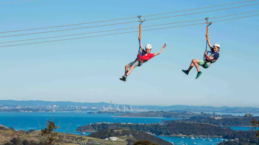 Two young people waving from the Waiheke zipline with Hauraki Gulf in the background