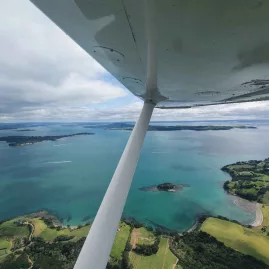 Aerial view of Waiheke Island and the Hauraki Gulf from a scenic flight, with the wing of a light aircraft visible overhead.