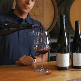 Person pouring red wine into a glass at a Waiheke Island vineyard, with bottles and barrels in the background during a premium Island Aviation wine and dining tour