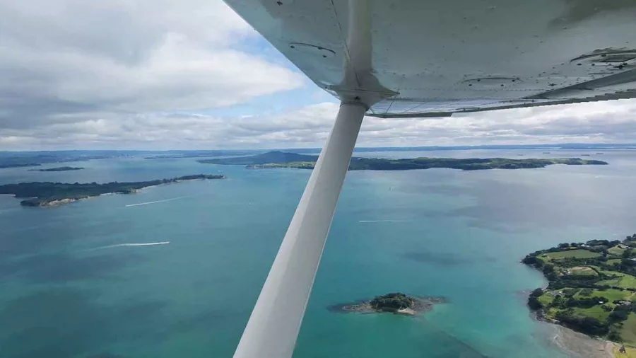 Aerial view of Waiheke Island and the Hauraki Gulf from a scenic flight, with the wing of a light aircraft visible overhead.