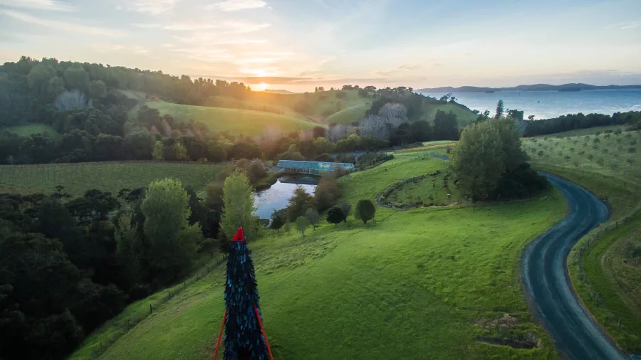 Aerial view of Brick Bay landscape in Snells Beach with green hills, sculpture, pond, winding road, and sunset sky