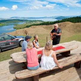 Māori guide playing guitar for visitors with sweeping views over Okawa Bay and Lake Rotoiti.