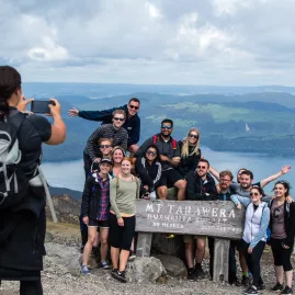 Group of hikers posing for a photo at the Mt Tarawera summit sign with Lake Tarawera in the background. Caption: