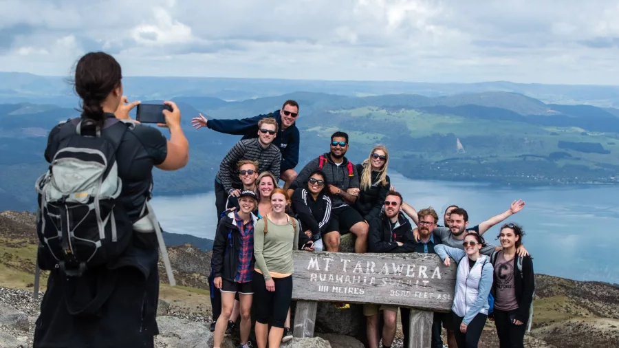 Group of hikers posing for a photo at the Mt Tarawera summit sign with Lake Tarawera in the background. Caption: