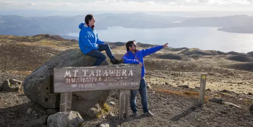 Two people beside the Mount Tarawera summit sign with sweeping views over Lake Tarawera.