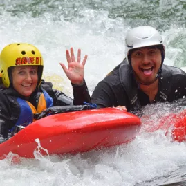 Two people sledging together through rapids, smiling and waving at the camera.