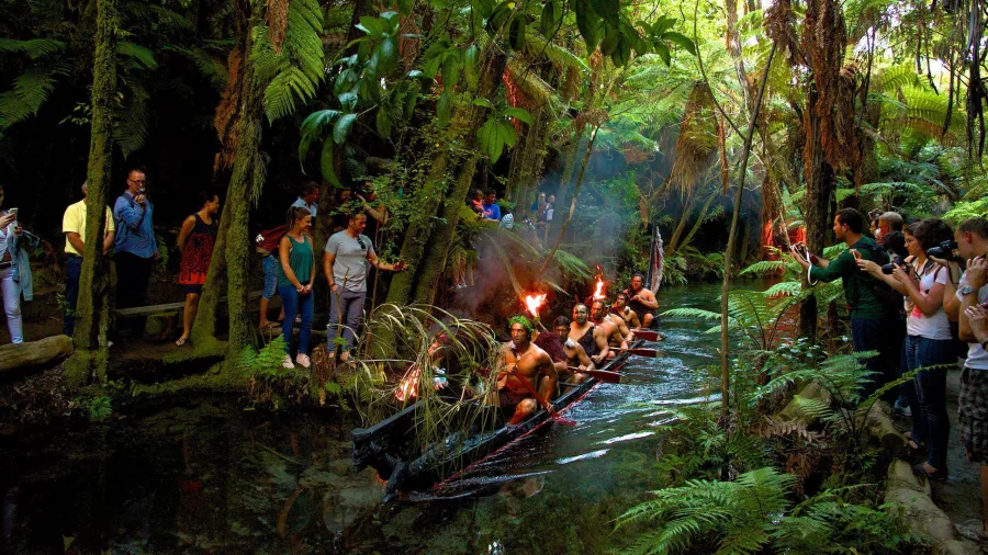 Māori warriors paddling a waka through native bush as visitors watch at Mitai Māori Village, Rotorua.